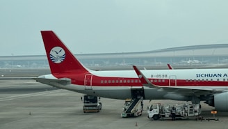 An airplane with the logo of Sichuan Airlines is parked on the tarmac of an airport. The aircraft's tail and part of its fuselage are visible, along with several service vehicles nearby. The background shows a large terminal building and a hazy sky.