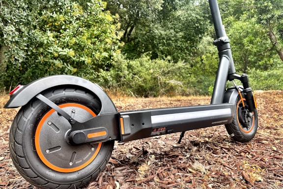 A modern electric scooter parked on a path with a backdrop of lush greenery. The close-up focuses on the rear wheel with orange accents and a sturdy black frame.