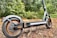 A modern electric scooter parked on a path with a backdrop of lush greenery. The close-up focuses on the rear wheel with orange accents and a sturdy black frame.