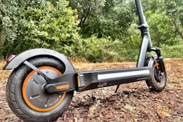 A modern electric scooter parked on a path with a backdrop of lush greenery. The close-up focuses on the rear wheel with orange accents and a sturdy black frame.