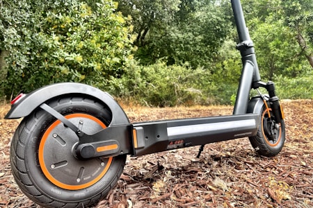 A modern electric scooter parked on a path with a backdrop of lush greenery. The close-up focuses on the rear wheel with orange accents and a sturdy black frame.