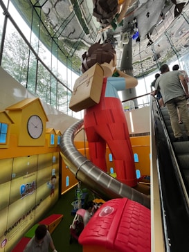 A large indoor playground with a giant figure resembling a child wearing a backpack, standing next to a large, twisting slide. The room has yellow walls with blue window graphics and a clock, and there are people using an escalator on the right. Additionally, there are toys and play structures on the green floor.