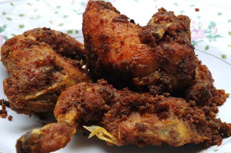 A close-up view of several pieces of deep-fried chicken arranged on a patterned plate. The chicken pieces are golden brown and crispy, with visible spices and crunchy texture on the surface. The plate shows a floral design around its edges.