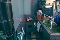 A narrow alley with brick walls lined with various objects including clothes hanging on a rack. A neon sign with the word 'SHELTER' is illuminated above a door. There are reflective surfaces and plants on a ledge, along with a blackboard and some scattered boxes.