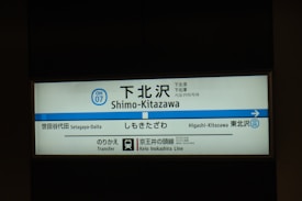 A subway or train station sign with the name Shimo-Kitazawa written in multiple languages, including Japanese, English, and Korean. The sign indicates an adjacent station, Higashi-Kitazawa, and mentions connections to the Keio Inokashira Line. The background is mostly shaded, highlighting the illuminated sign.