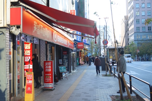 A bustling urban street scene featuring a row of shops with bright signage, including one with red and white text. Several people are walking along the sidewalk, some wearing jackets, indicating cooler weather. A few trees line the walkway, and a street runs parallel to the shops with a vehicle in the distance.