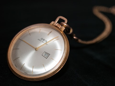A vintage round pocket watch with a gold casing and a silver dial, showing the date 23 in a small square window. The watch is resting on a dark surface, and a chain is visible connected to the watch's top.