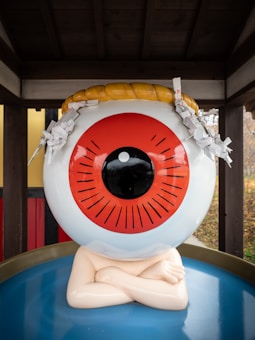 A large artistic sculpture in the shape of a human eye is displayed. The eye is predominantly white with a red iris and a black pupil. A decorative rope is placed on top and features paper ornaments attached. The sculpture is situated against a wooden backdrop with visible red and yellow panels.