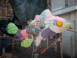 A large fish sculpture made from various colorful and patterned materials, including plastic bottles, fabric scraps, and other recycled items. The sculpture is placed outdoors, with a mesh fence and construction materials in the background.