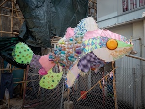 A large fish sculpture made from various colorful and patterned materials, including plastic bottles, fabric scraps, and other recycled items. The sculpture is placed outdoors, with a mesh fence and construction materials in the background.