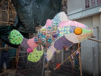A large fish sculpture made from various colorful and patterned materials, including plastic bottles, fabric scraps, and other recycled items. The sculpture is placed outdoors, with a mesh fence and construction materials in the background.