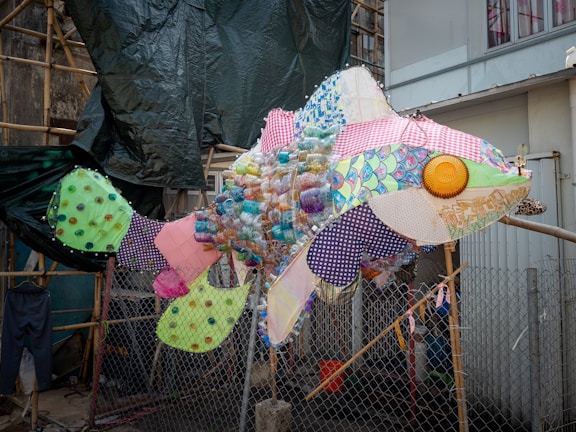 A large fish sculpture made from various colorful and patterned materials, including plastic bottles, fabric scraps, and other recycled items. The sculpture is placed outdoors, with a mesh fence and construction materials in the background.