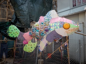 A large fish sculpture made from various colorful and patterned materials, including plastic bottles, fabric scraps, and other recycled items. The sculpture is placed outdoors, with a mesh fence and construction materials in the background.