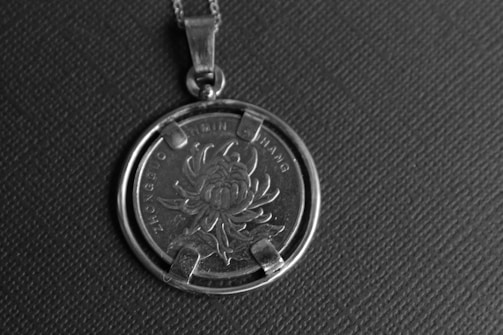 A close-up shot of a pendant featuring a coin with intricate floral designs, likely a chrysanthemum. The coin is encased in a metallic frame and suspended from a simple chain, set against a textured, dark background.