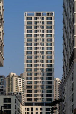A tall, modern apartment building with a grid-like design stands prominently between other similar high-rise structures. The sky is clear and blue, and the buildings feature large windows. There are neighboring buildings visible on either side, creating an urban canyon effect.