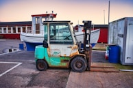 A weathered forklift is positioned in the foreground on an industrial dock. Behind it, a white fishing boat with a red cabin is visible. The surroundings include a red building and various industrial elements such as barrels and containers. The scene captures a rustic, maritime atmosphere.