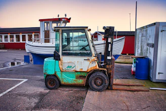 A weathered forklift is positioned in the foreground on an industrial dock. Behind it, a white fishing boat with a red cabin is visible. The surroundings include a red building and various industrial elements such as barrels and containers. The scene captures a rustic, maritime atmosphere.