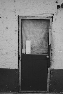A weathered, dark-colored door with a textured glass window set in an aged, stucco wall. The door appears old and slightly worn, with a small rectangular piece of material inside the window. The surrounding wall has a rough surface with visible patches and marks.