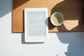 An e-reader displaying Chinese text is placed on a wooden surface, next to a small cup filled with a light-colored beverage. The scene is lit by natural sunlight, casting soft shadows and creating a calm atmosphere.