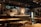 A cozy and warmly lit bar or restaurant interior featuring a long counter with multiple taps and some bottles displayed. The walls are decorated with various certificates, posters, and framed items. Tables and chairs are neatly arranged in the foreground, creating an inviting atmosphere.