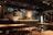 A cozy and warmly lit bar or restaurant interior featuring a long counter with multiple taps and some bottles displayed. The walls are decorated with various certificates, posters, and framed items. Tables and chairs are neatly arranged in the foreground, creating an inviting atmosphere.