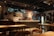 A cozy and warmly lit bar or restaurant interior featuring a long counter with multiple taps and some bottles displayed. The walls are decorated with various certificates, posters, and framed items. Tables and chairs are neatly arranged in the foreground, creating an inviting atmosphere.