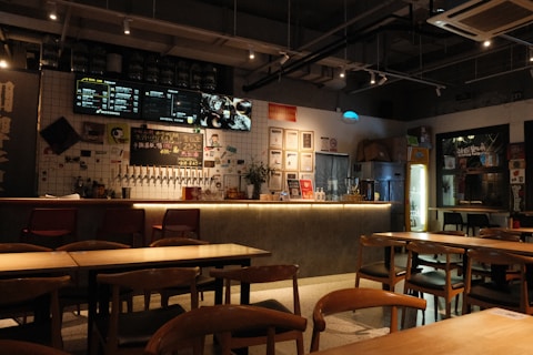 A cozy and warmly lit bar or restaurant interior featuring a long counter with multiple taps and some bottles displayed. The walls are decorated with various certificates, posters, and framed items. Tables and chairs are neatly arranged in the foreground, creating an inviting atmosphere.