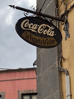 A vintage-looking Coca-Cola sign with the text 'La Vegueta de Colón' is mounted on a stone wall. The sign is oval-shaped, featuring classic Coca-Cola script in white against a black background. The wall has a mix of yellow and gray tones, with some electrical conduits visible. In the background, the top of a pink building with windows and a cloudy sky are visible.