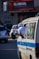 A person in a white outfit leans out of a window of a minibus in a busy urban street. Several cars are visible in the background, and there is a sign for an insurance company with red and white accents. Shadows and sunlight create a dynamic play of light on the scene.
