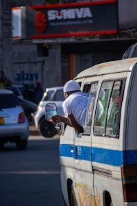 A person in a white outfit leans out of a window of a minibus in a busy urban street. Several cars are visible in the background, and there is a sign for an insurance company with red and white accents. Shadows and sunlight create a dynamic play of light on the scene.