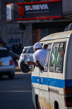 A person in a white outfit leans out of a window of a minibus in a busy urban street. Several cars are visible in the background, and there is a sign for an insurance company with red and white accents. Shadows and sunlight create a dynamic play of light on the scene.