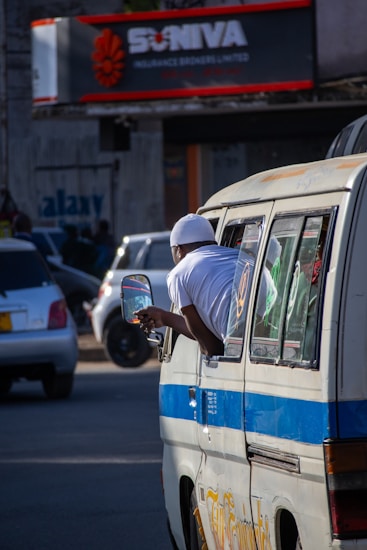 A person in a white outfit leans out of a window of a minibus in a busy urban street. Several cars are visible in the background, and there is a sign for an insurance company with red and white accents. Shadows and sunlight create a dynamic play of light on the scene.