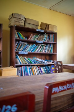 A wooden bookshelf filled with various books categorized by subjects like Physics, Mathematics, and Geography. Boxes labeled with text are placed on top of the shelf. The room has yellow walls and wooden furniture, suggesting it may be a classroom or library setting.