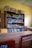 A wooden bookshelf filled with various books categorized by subjects like Physics, Mathematics, and Geography. Boxes labeled with text are placed on top of the shelf. The room has yellow walls and wooden furniture, suggesting it may be a classroom or library setting.