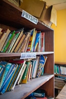 A wooden bookshelf filled with various textbooks and educational materials. Labels for 'Physics' and 'Mathematics' are seen on the shelves, indicating the subjects of the books. Some books are neatly arranged while others are leaning or stacked at angles. On top of the shelf is an open and slightly worn cardboard box.