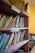A wooden bookshelf filled with various textbooks and educational materials. Labels for 'Physics' and 'Mathematics' are seen on the shelves, indicating the subjects of the books. Some books are neatly arranged while others are leaning or stacked at angles. On top of the shelf is an open and slightly worn cardboard box.