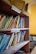A wooden bookshelf filled with various textbooks and educational materials. Labels for 'Physics' and 'Mathematics' are seen on the shelves, indicating the subjects of the books. Some books are neatly arranged while others are leaning or stacked at angles. On top of the shelf is an open and slightly worn cardboard box.