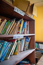 A wooden bookshelf filled with various textbooks and educational materials. Labels for 'Physics' and 'Mathematics' are seen on the shelves, indicating the subjects of the books. Some books are neatly arranged while others are leaning or stacked at angles. On top of the shelf is an open and slightly worn cardboard box.