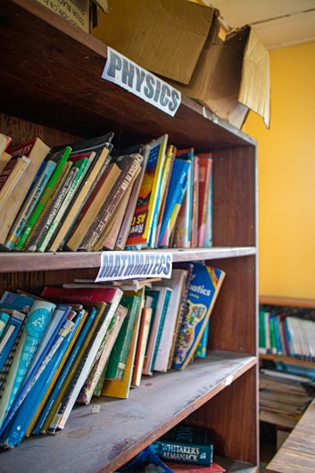 A wooden bookshelf filled with various textbooks and educational materials. Labels for 'Physics' and 'Mathematics' are seen on the shelves, indicating the subjects of the books. Some books are neatly arranged while others are leaning or stacked at angles. On top of the shelf is an open and slightly worn cardboard box.