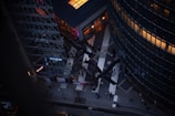 An aerial view of an urban landscape with modern architecture, including a prominent IMAX theater sign on a glass building. People are walking in a plaza-like area with geometric patterns on the ground, flanked by two high-rise buildings with reflective glass windows. The lighting is dim, suggesting dusk or early evening.