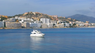 A white yacht floats on calm blue water in a coastal area with a backdrop of numerous multi-story buildings, and a hillside dotted with houses. A mountain range can be seen in the distance under a clear sky.