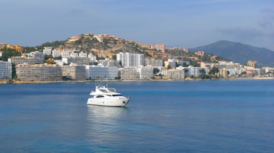 A white yacht floats on calm blue water in a coastal area with a backdrop of numerous multi-story buildings, and a hillside dotted with houses. A mountain range can be seen in the distance under a clear sky.