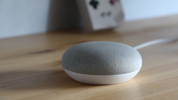 A small, rounded smart speaker with a mesh fabric covering sits on a wooden surface. In the background, there is a blurred object resembling a retro gaming console leaning against the wall.
