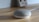 A small, rounded smart speaker with a mesh fabric covering sits on a wooden surface. In the background, there is a blurred object resembling a retro gaming console leaning against the wall.