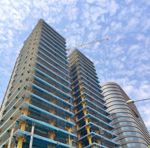 Tall buildings under construction with visible scaffolding and a construction crane. The sky is bright blue and partly cloudy, providing a backdrop to the urban scene. Another completed, modern, curved building is visible to the right.
