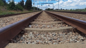 A straight railway track extends toward the horizon, surrounded by gravel on the ground. The sky is partially cloudy with some blue visible, and there are green trees and grass along the sides of the track. Overhead lines and metal poles are visible above the tracks.