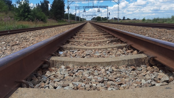 A straight railway track extends toward the horizon, surrounded by gravel on the ground. The sky is partially cloudy with some blue visible, and there are green trees and grass along the sides of the track. Overhead lines and metal poles are visible above the tracks.
