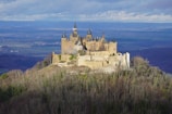A majestic castle sits atop a lush, green hill surrounded by a dense woodland. The structure features numerous spires and turrets, with a mix of light and darker stones. In the background, a vast, flat landscape stretches out to a distant horizon under a cloudy sky.