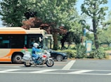 A motorcyclist wearing a white helmet and blue uniform rides a police motorcycle in front of an orange and cream city bus. The scene is set in an urban area with lush green trees and foliage in the background. A black car is partially visible behind the bus. The pavement is marked with white road crossing lines.