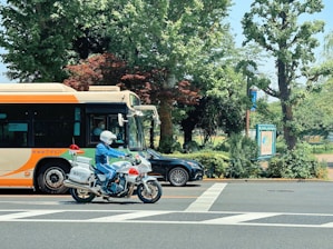 A motorcyclist wearing a white helmet and blue uniform rides a police motorcycle in front of an orange and cream city bus. The scene is set in an urban area with lush green trees and foliage in the background. A black car is partially visible behind the bus. The pavement is marked with white road crossing lines.